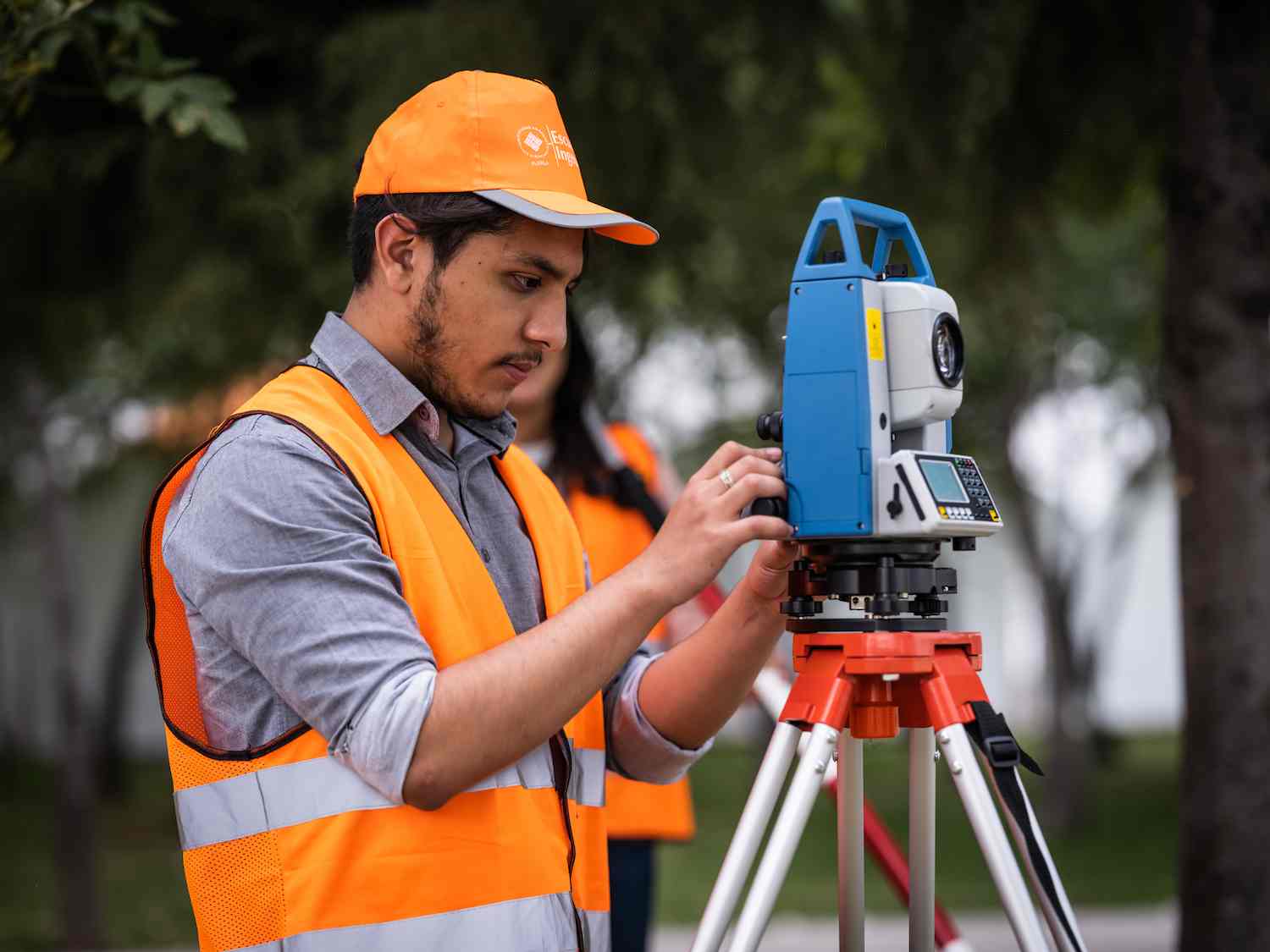Estudiante ajusta estación total de topografía sobre trípode durante práctica al aire libre con chaleco y gorra de seguridad.